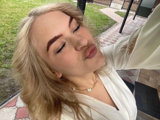 AlinaLemon with blonde hair, closed eyes, and pouty lips, wearing a white top and gold necklace, taking a selfie. Her eyebrows are thick and she's wearing small earrings.