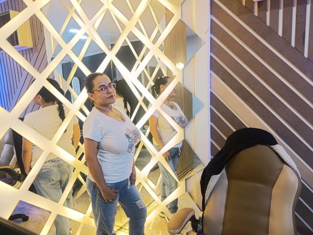 SaraParkers with glasses, white t-shirt, and blue jeans, standing in front of mirrored wall, showing reflection of her and another woman's ass in jeans. Chair and stairs nearby.