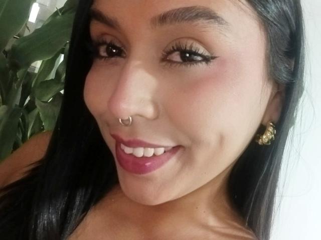 ZammySanz ha un viso carino, con capelli lunghi e scuri, un piercing al naso e un grande sorriso che mostra il rossetto rosa. Indossa orecchini dorati e ha l'eyeliner..