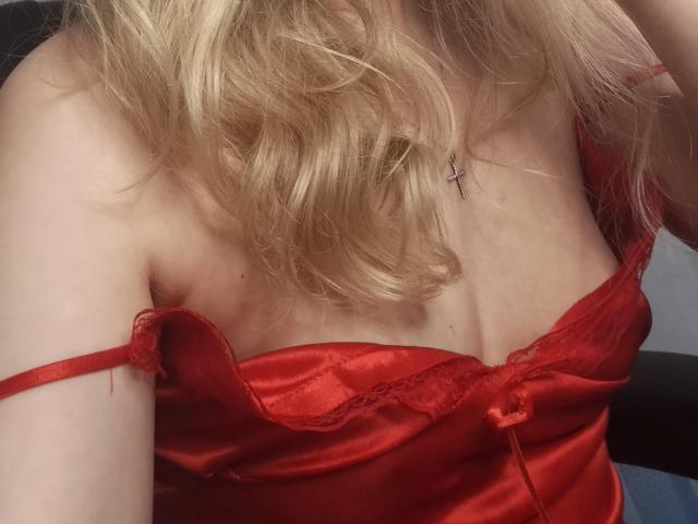 GladisBelle's red satin lingerie, showing her tits with lace trim, off-shoulder, blonde wavy hair, and a cross necklace, in a soft, intimate photo.
