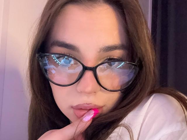 RoseleeVandiest with reflective glasses, pink nails, and finger to lips, has brown hair and a cute face, wearing a white shirt, in a softly lit room.