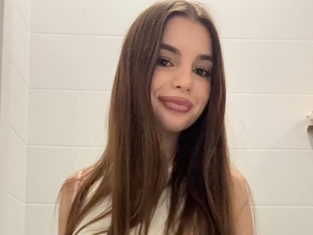 RoseleeVandiest with long brown hair, smiling with pink lipstick, white tank top, and bare shoulders, in a bathroom with white tiled walls.