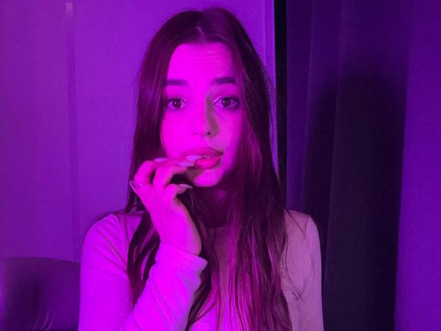 RoseleeVandiest with pink-purple lit face, long brown hair, white long-sleeve shirt, hand near pink-lit lips, looking at camera. Soft light, serious expression.