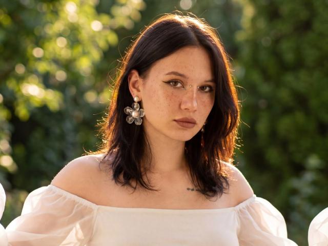 AlicePertim with freckled face, dark wavy hair, white off-shoulder dress, puffy sleeves, flower earrings, and a small tattoo above her chest, looking serious in sunlight.