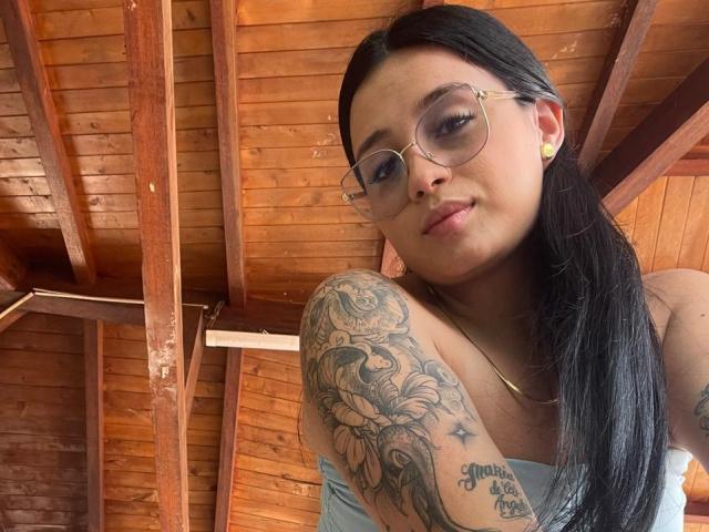 AnnyBentleii with long black hair, glasses, and tattoos on her right shoulder, looking down, wearing a light blue top, wooden ceiling in background, small gold earrings.