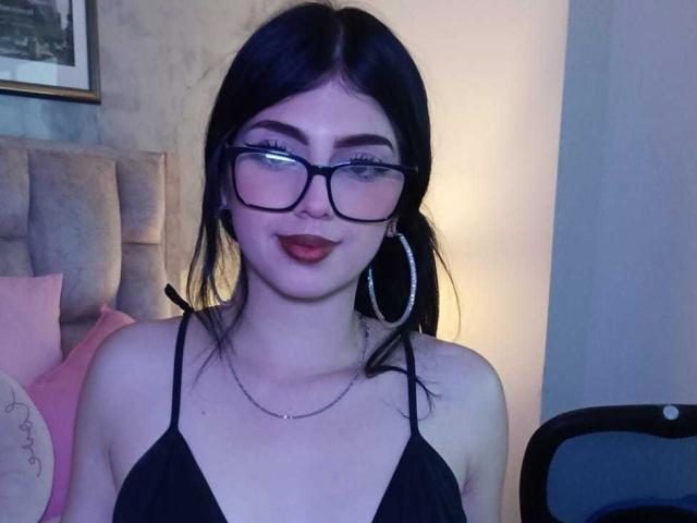 IsabellaaRossi with glasses, black hair, red lipstick, hoop earrings, black halter top, necklace, smiling, showing tits, fair skin, sitting in dimly lit room, holding phone.