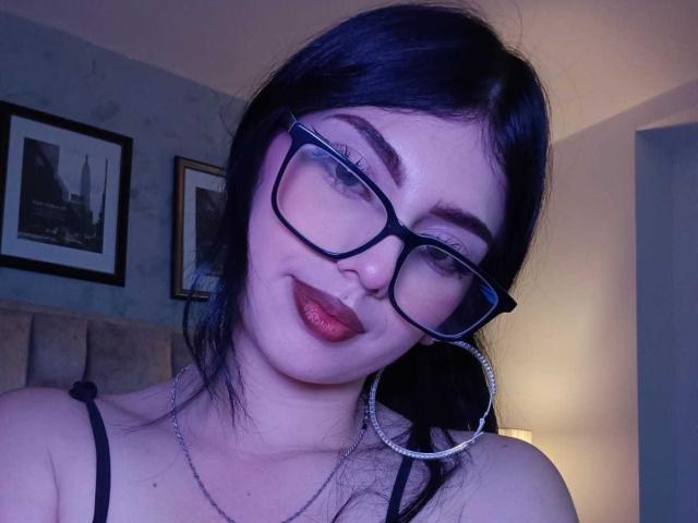 IsabellaaRossi with dark blue hair, black glasses, red lipstick, and large silver hoop earrings, smiling slightly, showing her upper chest and a thin black strap.