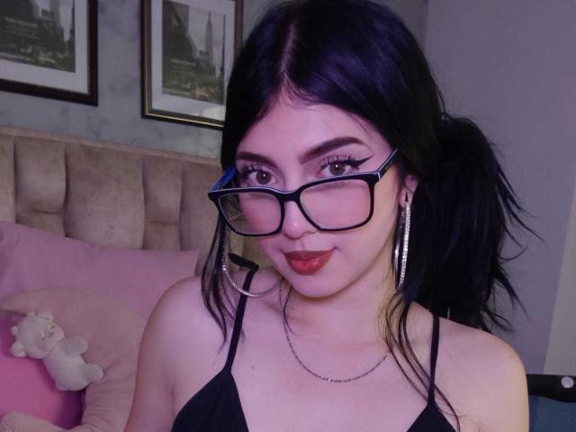 IsabellaaRossi with black pigtails, glasses, hoop earrings, and red lipstick, showing off her small tits in a black top, sitting on a pink bed with a stuffed toy.