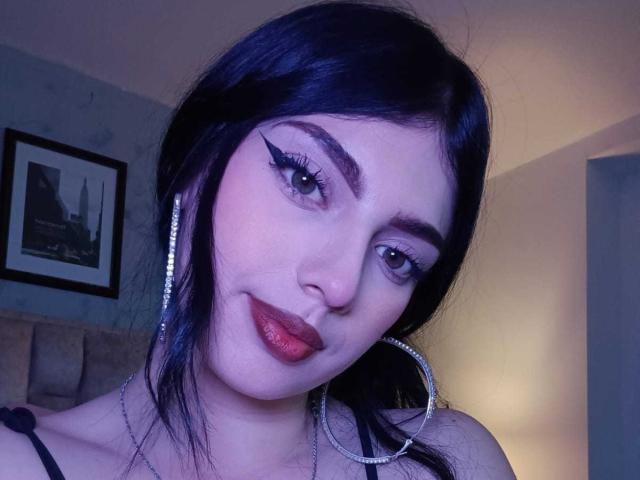 IsabellaaRossi has dark blue hair, sharp winged eyeliner, glittery red lipstick, and a silver hoop earring, with a slight smirk, wearing a black strap top.