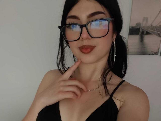 IsabellaaRossi with black glasses, red lipstick, and a finger to her lips, wearing a black top, showing her tits, and a necklace, against a plain background.