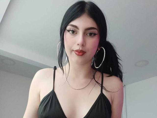 IsabellaaRossi with black hair, red lipstick, hoop earrings, chain necklace, and black dress with thin straps, showing her tits and small chest, in a selfie.