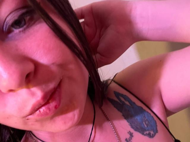 GraceRedMiller with a blue wolf tattoo on her shoulder, puffy lips, and a nose ring, touching her brown hair, wearing a black strap, under pink lighting.