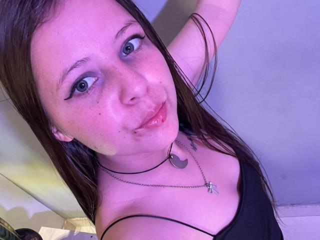 GraceRedMiller with long brown hair, blue eyes, and pinkish skin, wearing a black top, has a small nose piercing, and is smiling with her arm raised, showing a necklace.