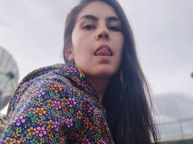 SweetShanny, con cabello largo y oscuro, la lengua afuera, vistiendo una sudadera con capucha de colores y estampado floral. Su rostro está centrado, ligeramente inclinado hacia arriba, con una expresión relajada y juguetona..