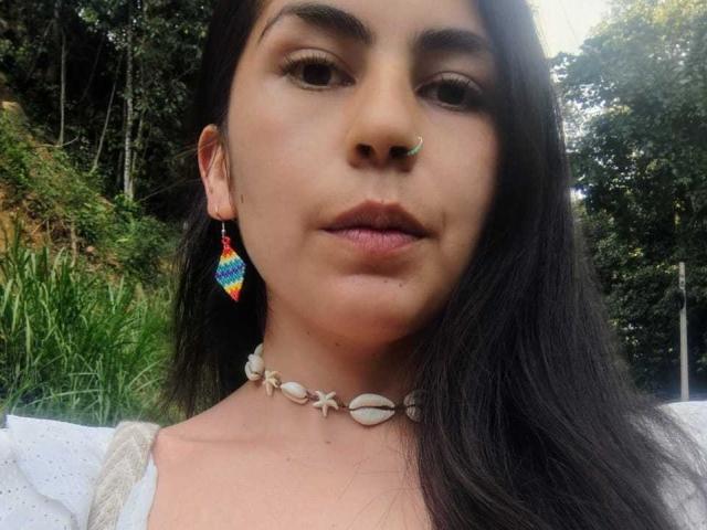 SweetShanny has dark hair, a nose ring, rainbow earrings, and a shell choker, with subtle makeup and a neutral expression, showing her face and upper chest in a selfie.