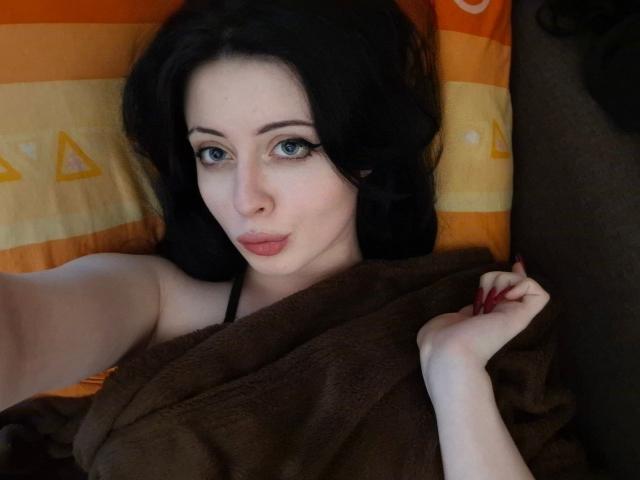 DanielleZiego with black hair, blue eyes, and pink lips, lying in bed, covered by a brown blanket, taking a selfie, showing her tits, with red nails.