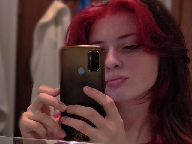 DanielleZiego with red and black hair, holding a brown phone, taking a mirror selfie with pink nails, slightly pouty lips, and a subtle smile.