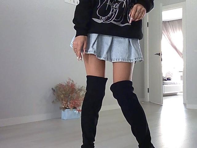 EmiliaMuse trägt einen schwarzen, übergroßen Pullover, einen hellblauen Jeansrock und schwarze, hohe Stiefel. Sie steht in einem Raum mit weißen Wänden und einer Tür..
