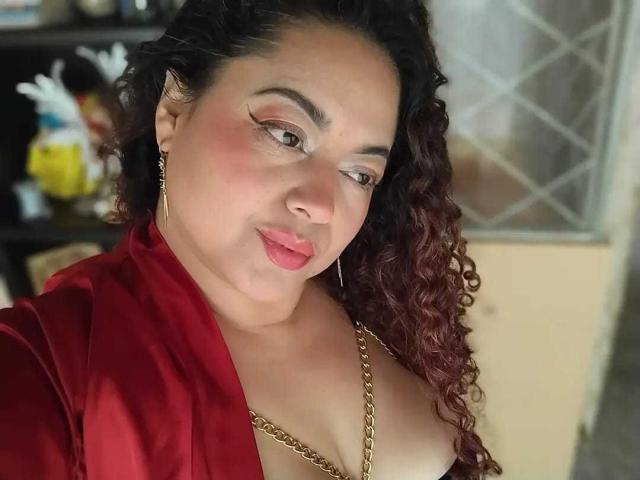 SophiLarusso with curly brown hair, red lipstick, and gold earrings, wearing a red satin robe, showing cleavage with a gold chain, has a slight smile, and pink eye makeup.