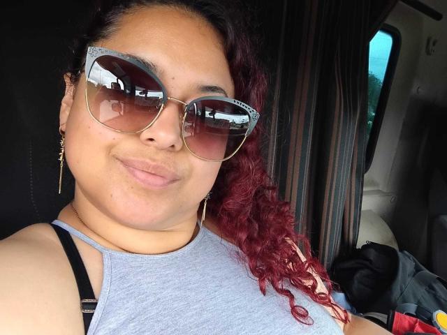 SophiLarusso with curly red hair, wearing large reflective sunglasses, grey tank top, and black straps, smiling softly, looking cute and relaxed. Her tits are small, and she's sexy.