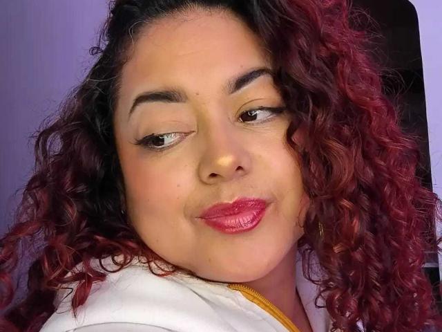 SophiLarusso has curly red hair, dark eyebrows, light skin, and glossy pink lips. She's wearing a white shirt with yellow trim, looking slightly to the side with a smirk.
