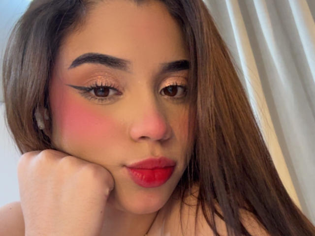 LunaVali has red lipstick, pink blush, and winged eyeliner, resting her chin on her hand with long brown hair, looking at the camera with brown eyes.