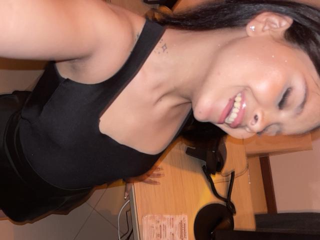 ChloeVixen with dark hair, smiling, wearing a black tank top, showing small tits, and a tattoo near her neck, leaning over a desk with headphones and a mask.
