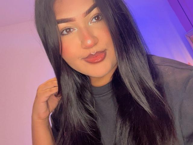 DojaAissha with long black hair, brown eyes, thick eyebrows, red lipstick, and a nose ring, touching her hair with a slight smile, wearing a black top.