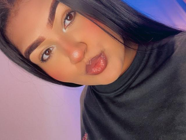DojaAissha has shiny brown eyes, full pink lips, and dark brown eyebrows. She's wearing a black turtleneck with long black hair. Her face is slightly tilted to the right.