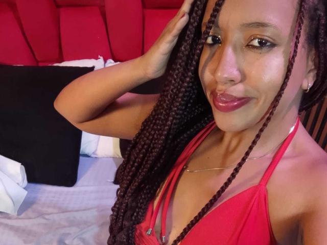 LilithAndAsmodeo with long braids, wearing a red halter top, smiling, showing her tits, hand in hair, red lipstick, small earrings, and a necklace, in a bedroom.