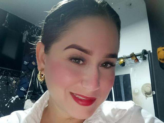 AlexaHotSeins has red lipstick, flushed cheeks, dark eyebrows, and is smiling with a white shirt on, wearing gold earrings, and has a nose ring, with dark hair in a bun.