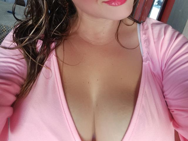 AlexaHotSeins with wet brown hair, pink zip-up shirt, and big, bare tits showing cleavage, wearing pink lipstick, upper chest and face partially visible.