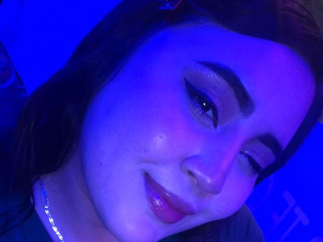 GraceYuly with blue-tinted face, winking, and smiling, has glossy pink lips, dark eyebrows, and subtle eyeshadow, wearing a necklace, with brown hair, in a low-light photo.