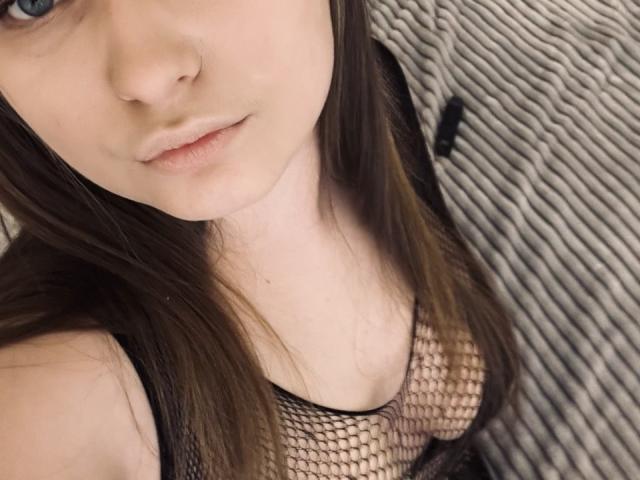 MagikPeachh with pale skin, brown hair, and blue eyes, wearing a black fishnet top that reveals her small tits, has subtle pink lips, and a slightly serious expression.