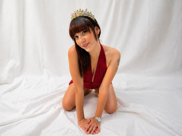 HanaJoud, with brown hair and a tiara, kneels on white cloth, wearing a red halter top, exposing her small tits and showing a tattoo on her right arm.