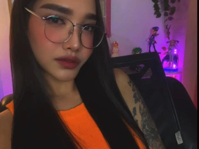 SooHotN2Sexy-hot with long black hair, glasses, orange top, and tattooed arm, sitting in a chair, looking at camera with pinkish-purple lighting in the background.
