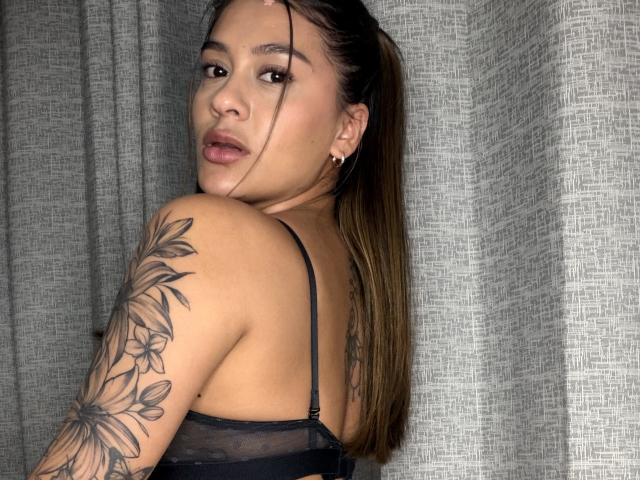 ThaliaAres with long brown hair in a ponytail, looking over her shoulder, wearing a black sheer top, showing a floral tattoo on her right arm, and small earrings.