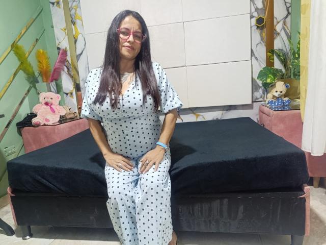 SaraParkers, with long dark hair and red glasses, sits on a black-covered bed in a white polka-dot dress, hands on her ass, looking slightly bored.