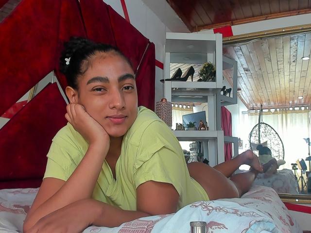 NatalyBlonder, lying on bed, green shirt, black hair bun, head resting on hand, cute smile, bare ass, white sheets, wooden ceiling, red wall, shelves with plants, daytime.