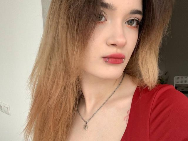 LianaKisses with light brown hair, red lipstick, and a nose piercing, wearing a red shirt, silver necklace, and a tattoo on her right chest. She has long eyelashes.