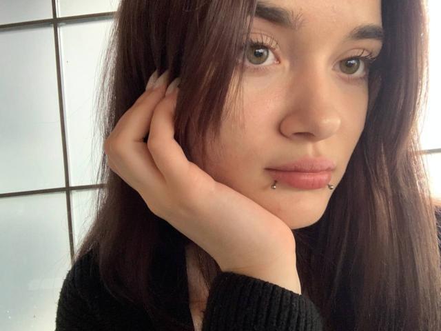 LianaKisses has long brown hair, green eyes, and a black lip piercing. She's holding her face with one hand, wearing a black sweater, with a slightly curious expression.