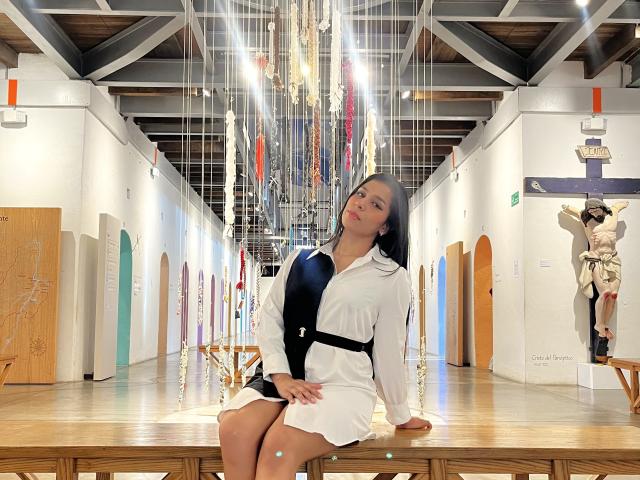 MelodyRoss, with long black hair, sits on a wooden table in a white dress with black accents, looking up with a slight smile. Her ass is partially visible.
