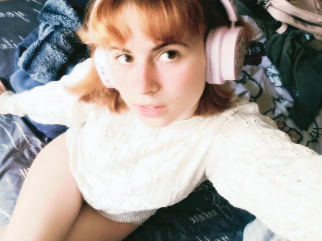 MireilleSpotwood with pink headphones, white knit sweater, and bare thighs, sitting on a bed, looking up with pale skin and red hair, soft expression.