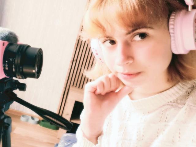 MireilleSpotwood with pink headphones, blonde hair, and white sweater, holding her chin, next to a camera on a tripod, looking at the camera.