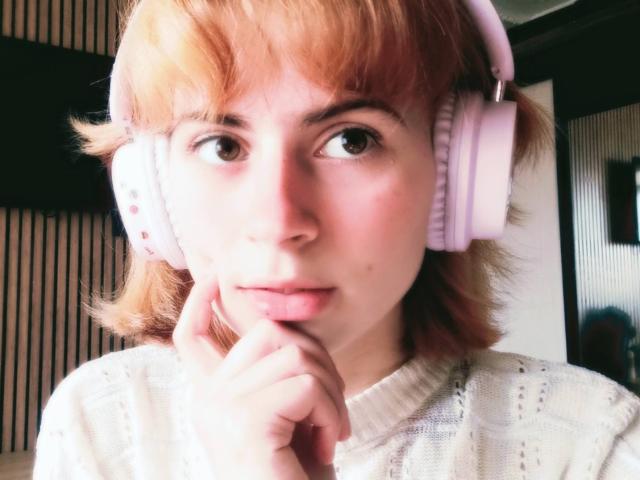 MireilleSpotwood has short, orange hair, wearing pink headphones, with her finger touching her chin, wearing a white, knitted sweater, with a slightly flushed, cute face.
