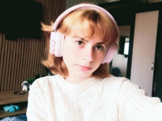 MireilleSpotwood with short, blonde hair, wearing pink headphones and a white, knit sweater, has a slight, pouty expression. Her tits are small, and her ass is not visible.