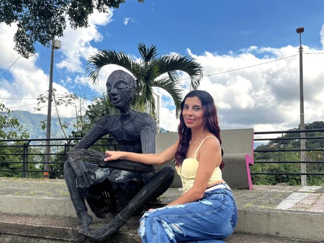 Carla Torres, con cabello oscuro, una camiseta amarilla y jeans azules desgastados, sentada al lado de una estatua negra y esculpida, con la mano sobre su rodilla, sonriendo a la cámara..