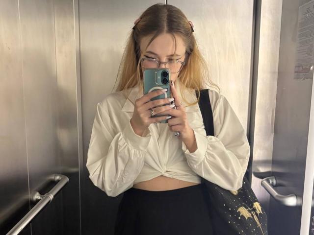 LunaBerryy with glasses, blonde hair in pigtails, white tied-up shirt, black skirt, holding blue phone in elevator, black bag with gold stars, mirror selfie, looking down.