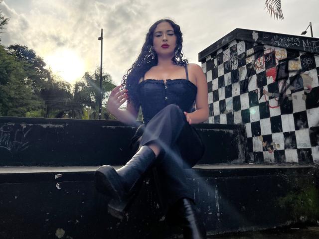 LilithStygian with curly black hair, dark red lipstick, and a strapless black dress, sitting on black steps, legs crossed, wearing black boots, against a graffiti-covered wall.