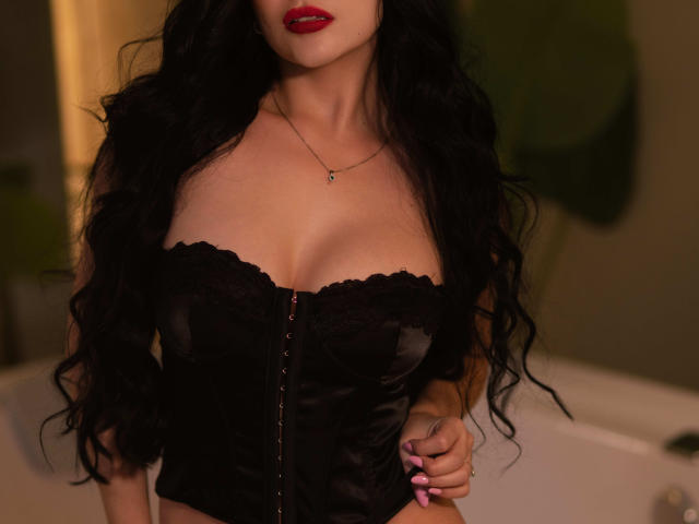 ManuelaOcampoz with long black hair, red lipstick, wearing a black lace-up corset, showing her large tits, hands resting on her hips, a delicate necklace, and pink nails.